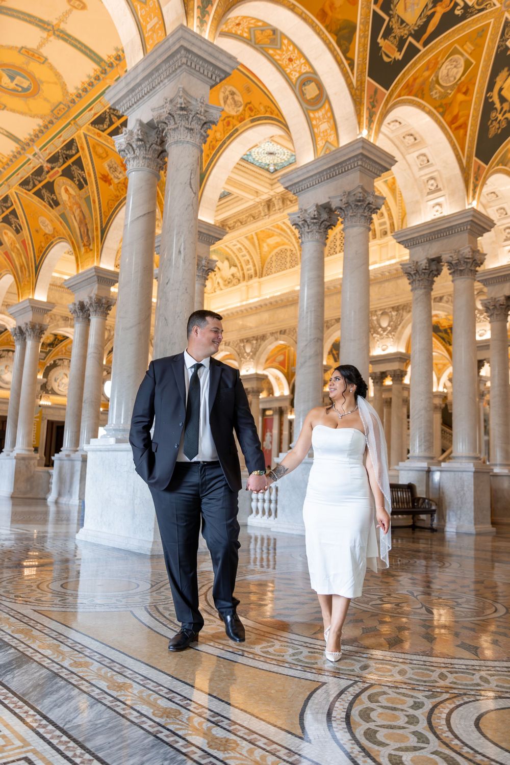 Wedding couple walks through ornate golden hallway with arched ceilings and columns.