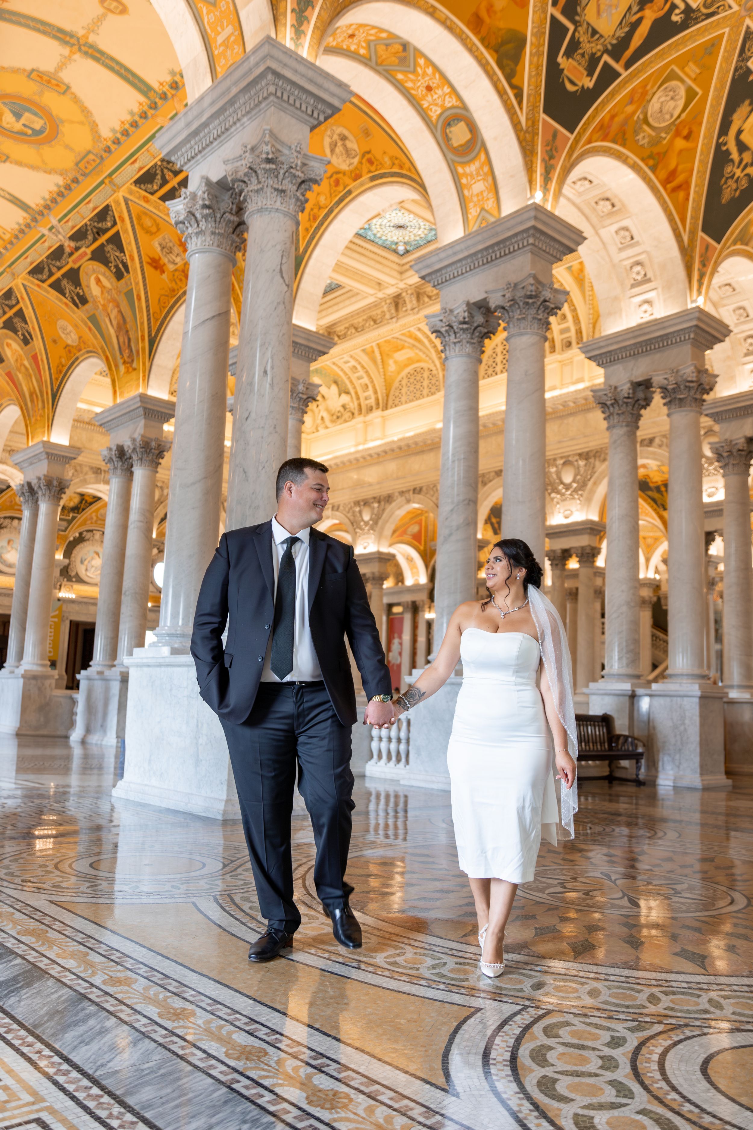 Wedding couple walks through ornate golden hallway with arched ceilings and columns.