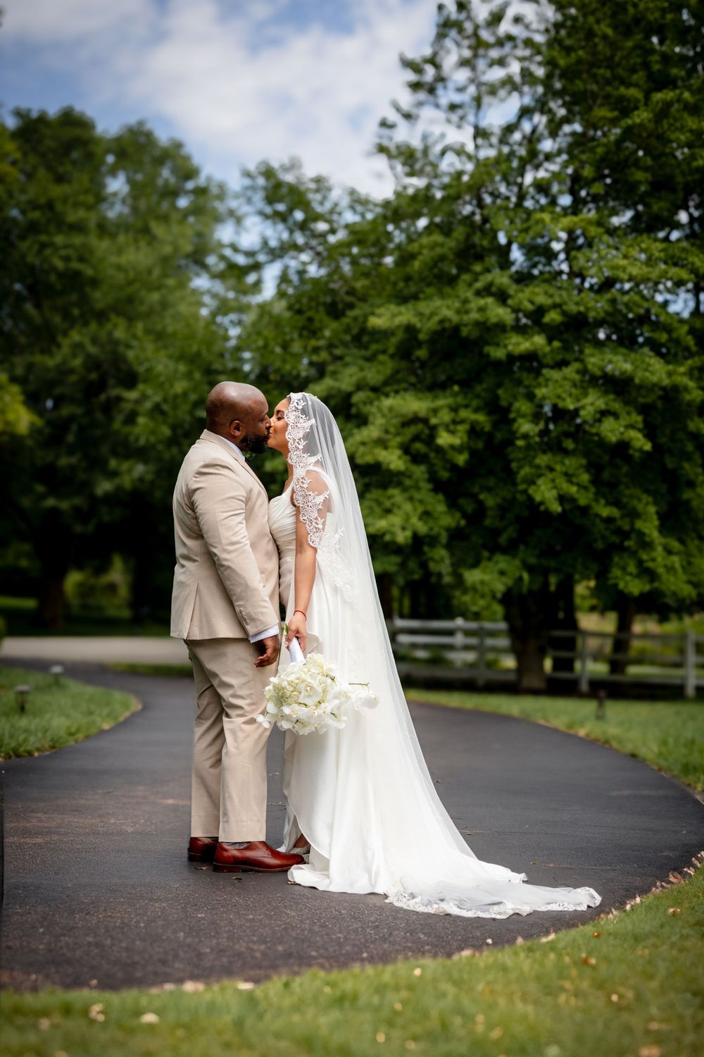 Romantic wedding couple sharing a tender moment on a tree-lined path in their formal attire.