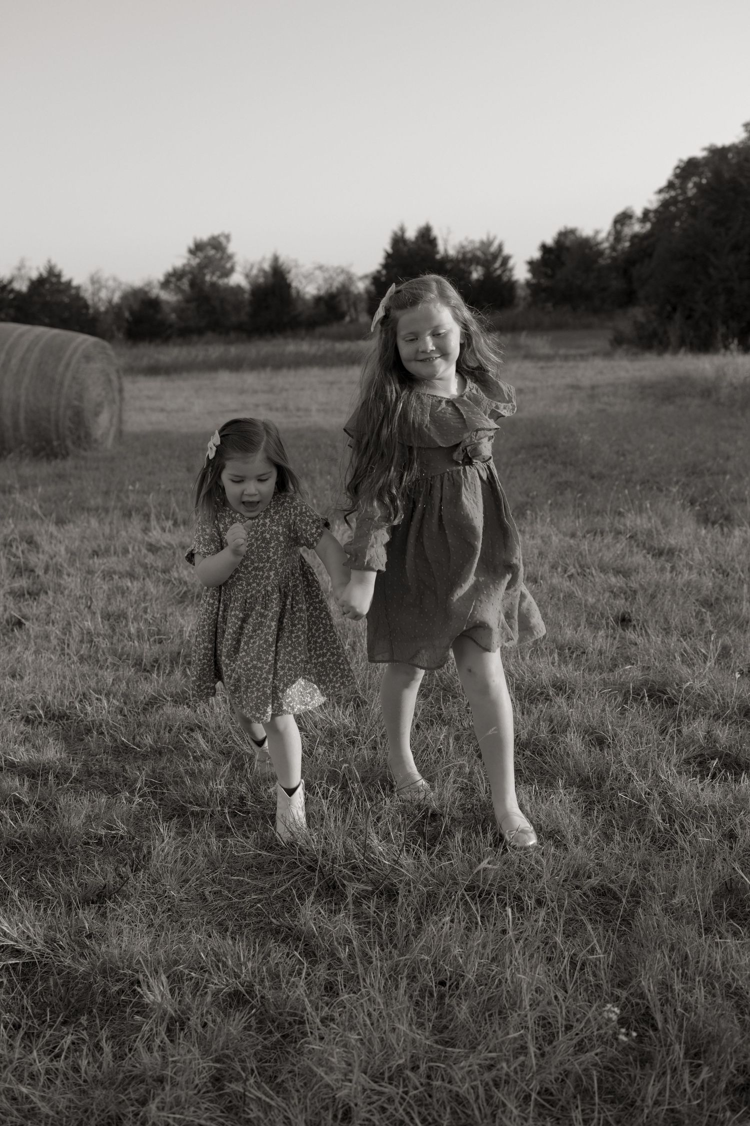Two children in dresses running and playing together in a grassy field at dusk.