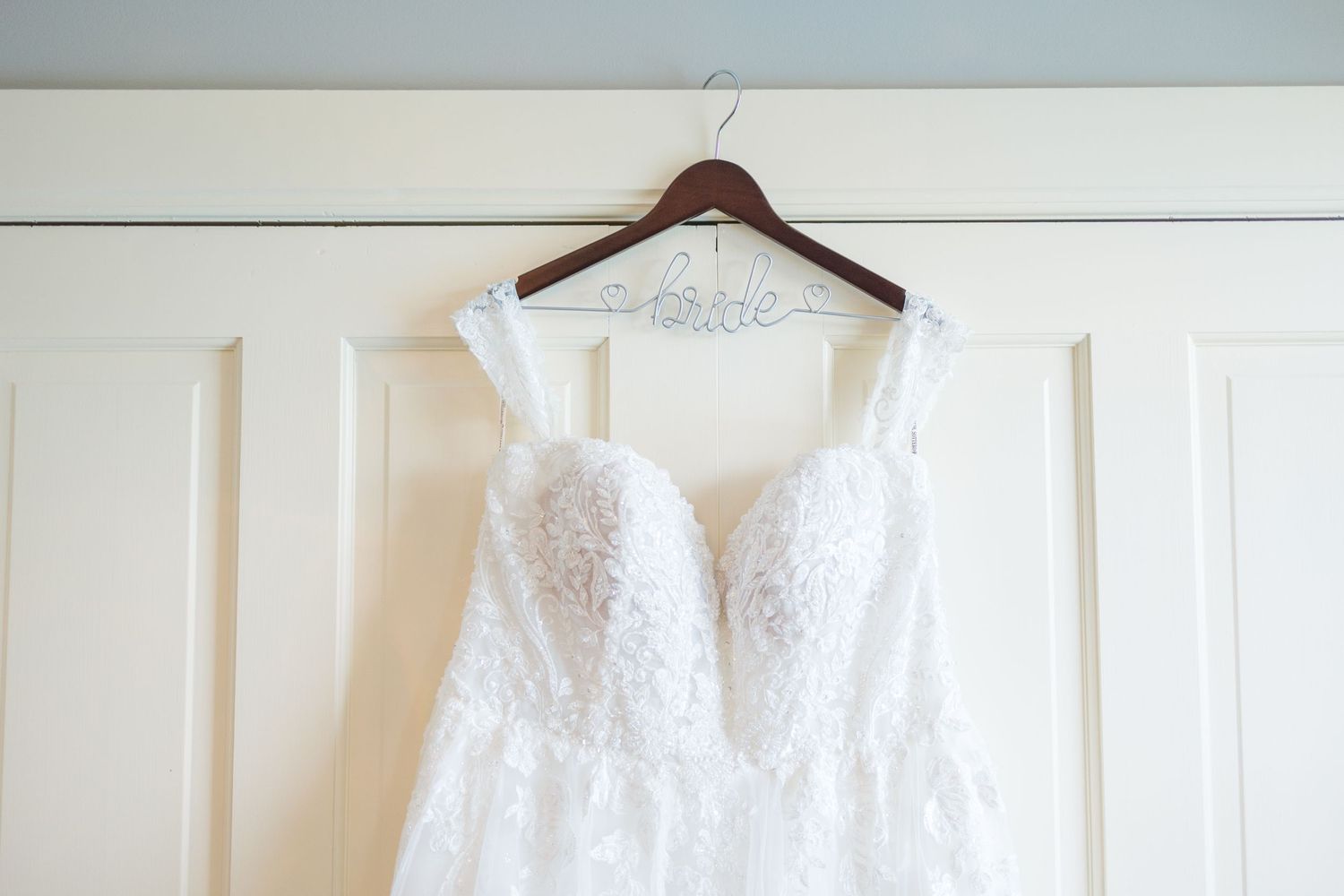 A white lace wedding dress hanging on a wooden hanger against a white wall.