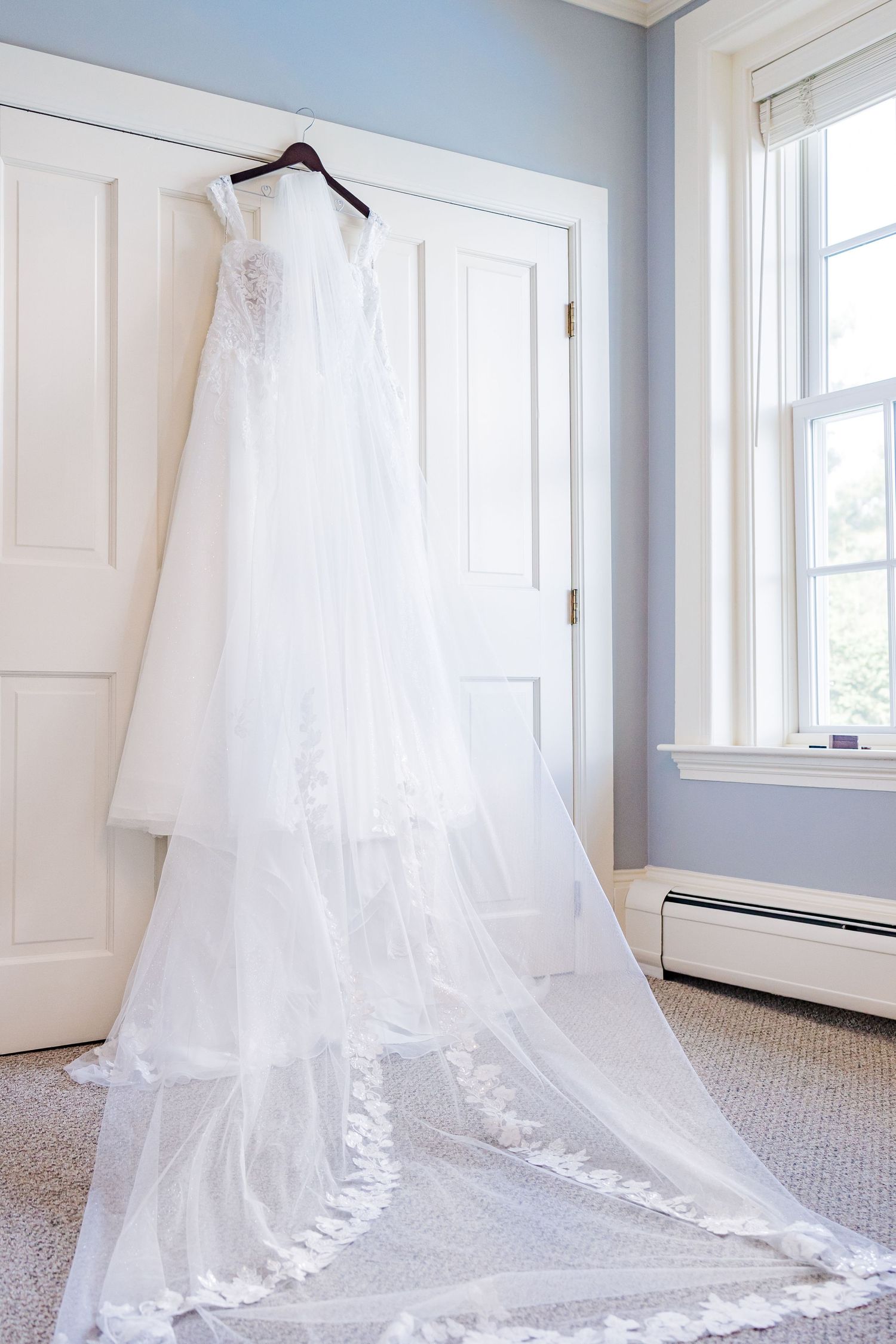 An elegant white lace wedding gown with long train displayed on a hanger against a white door.