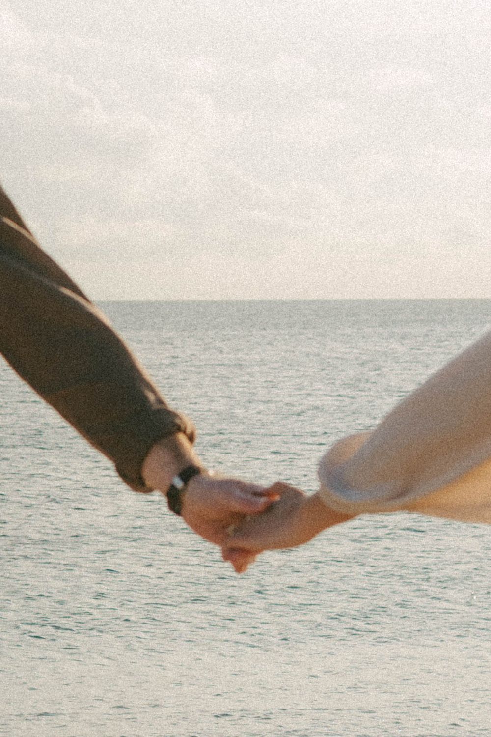 Close-up of two hands holding each other against an ocean backdrop at sunset, creating a romantic silhouette.