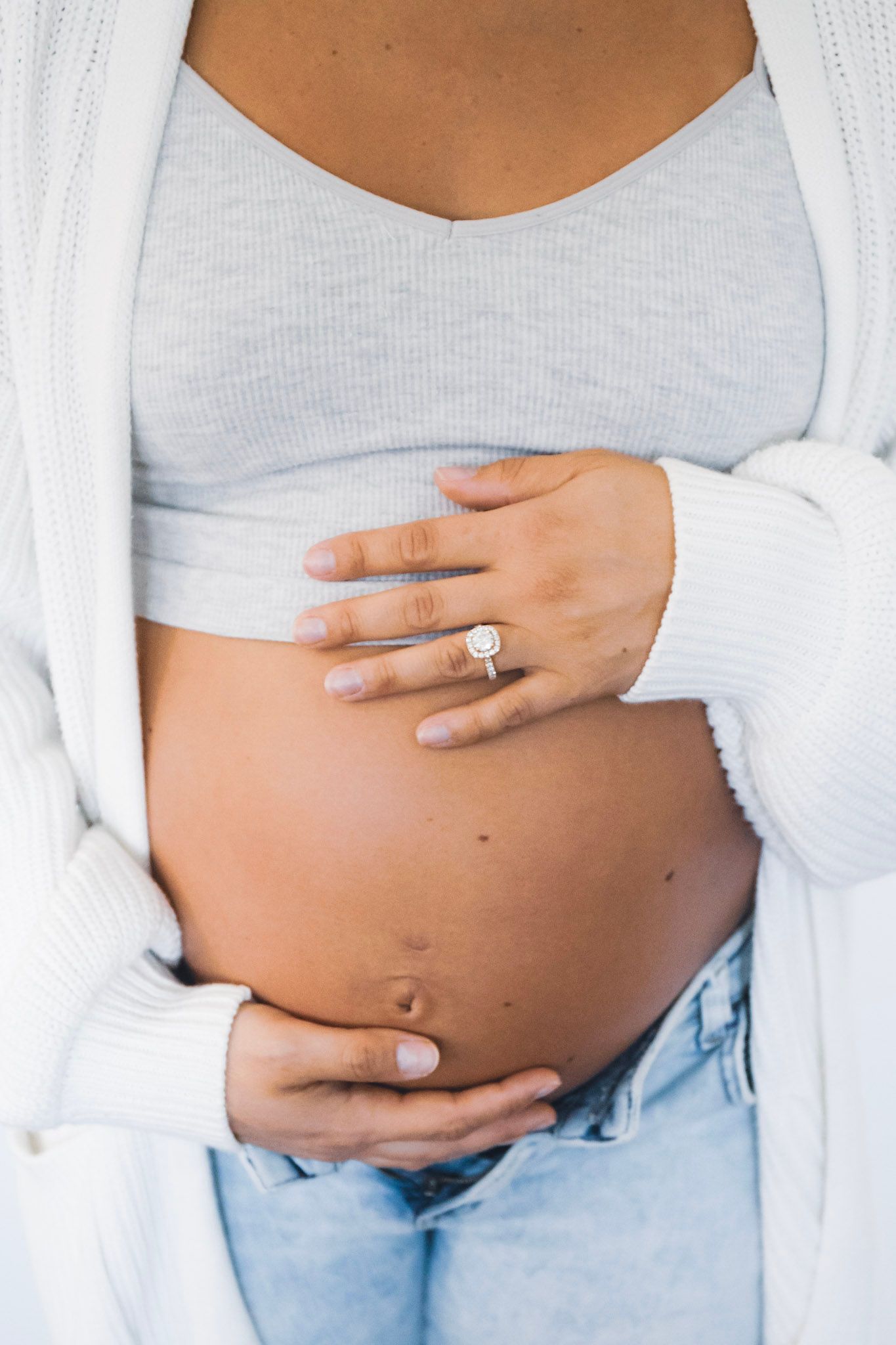 Pregnant woman in white cardigan holding her belly with engagement ring visible.