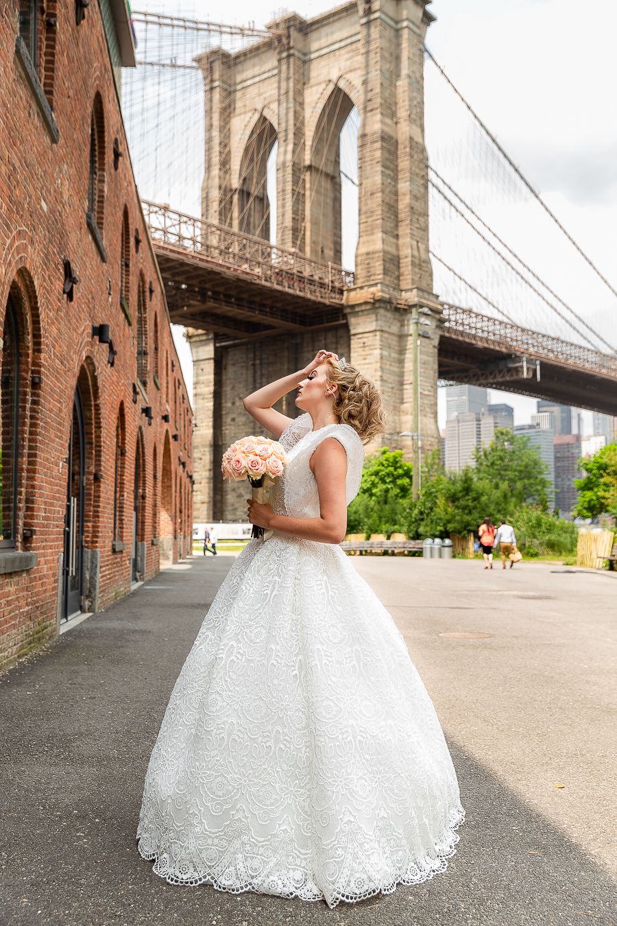Bride in white lace ball gown poses near Brooklyn Bridge with historic brick buildings.