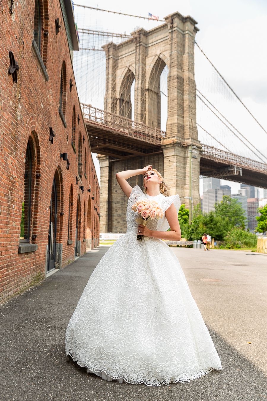 An elegant bridal portrait showcasing a white lace ball gown against the backdrop of the Brooklyn Bridge and brick buildings.