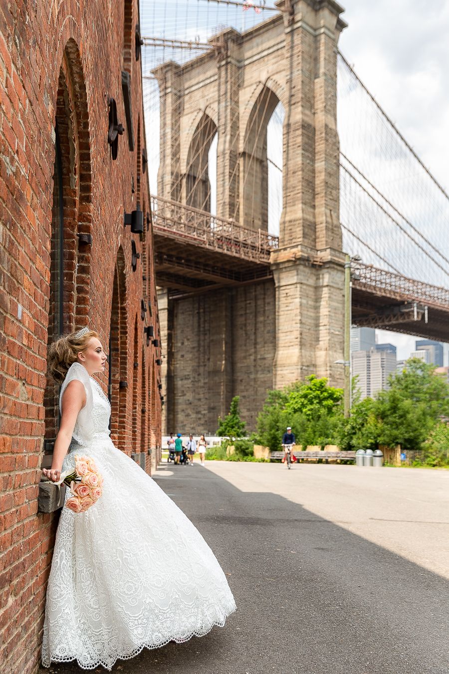 A classic wedding photo featuring a full lace gown against red brick walls and the Brooklyn Bridge.