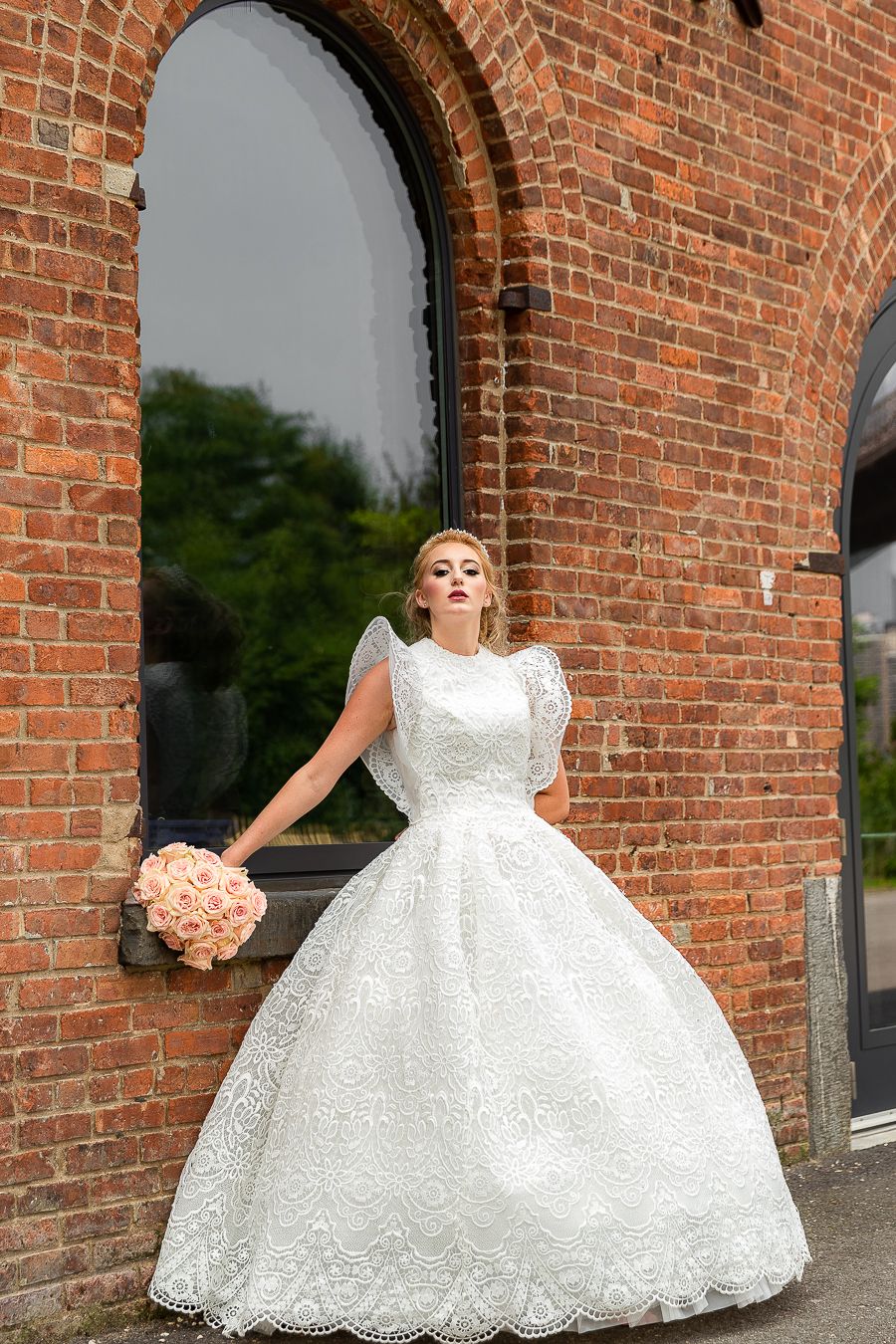 A dramatic bridal pose in a lace ball gown with peach roses against a brick archway.
