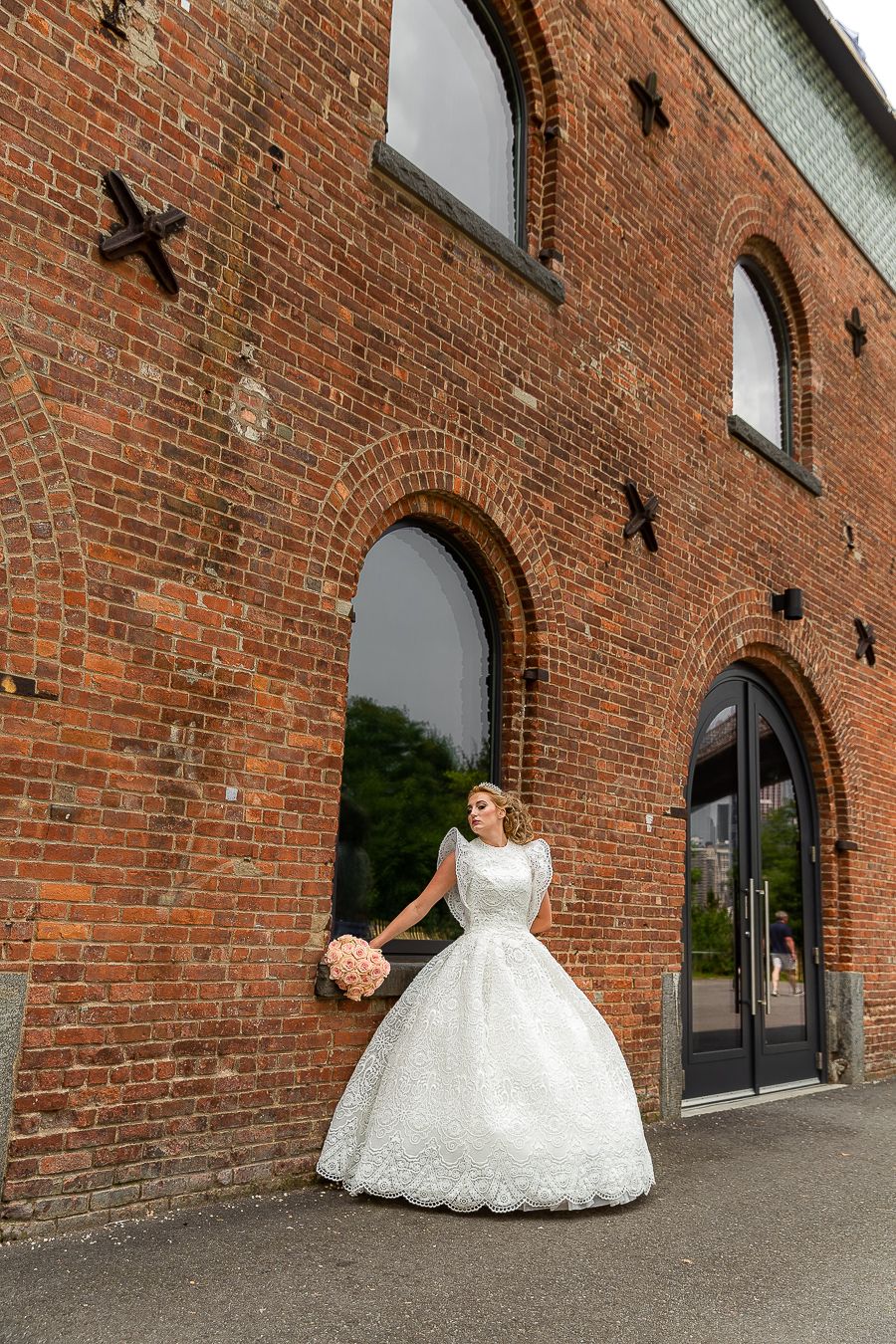 Elegant wedding gown with full lace skirt shown against rustic brick building with arched windows.