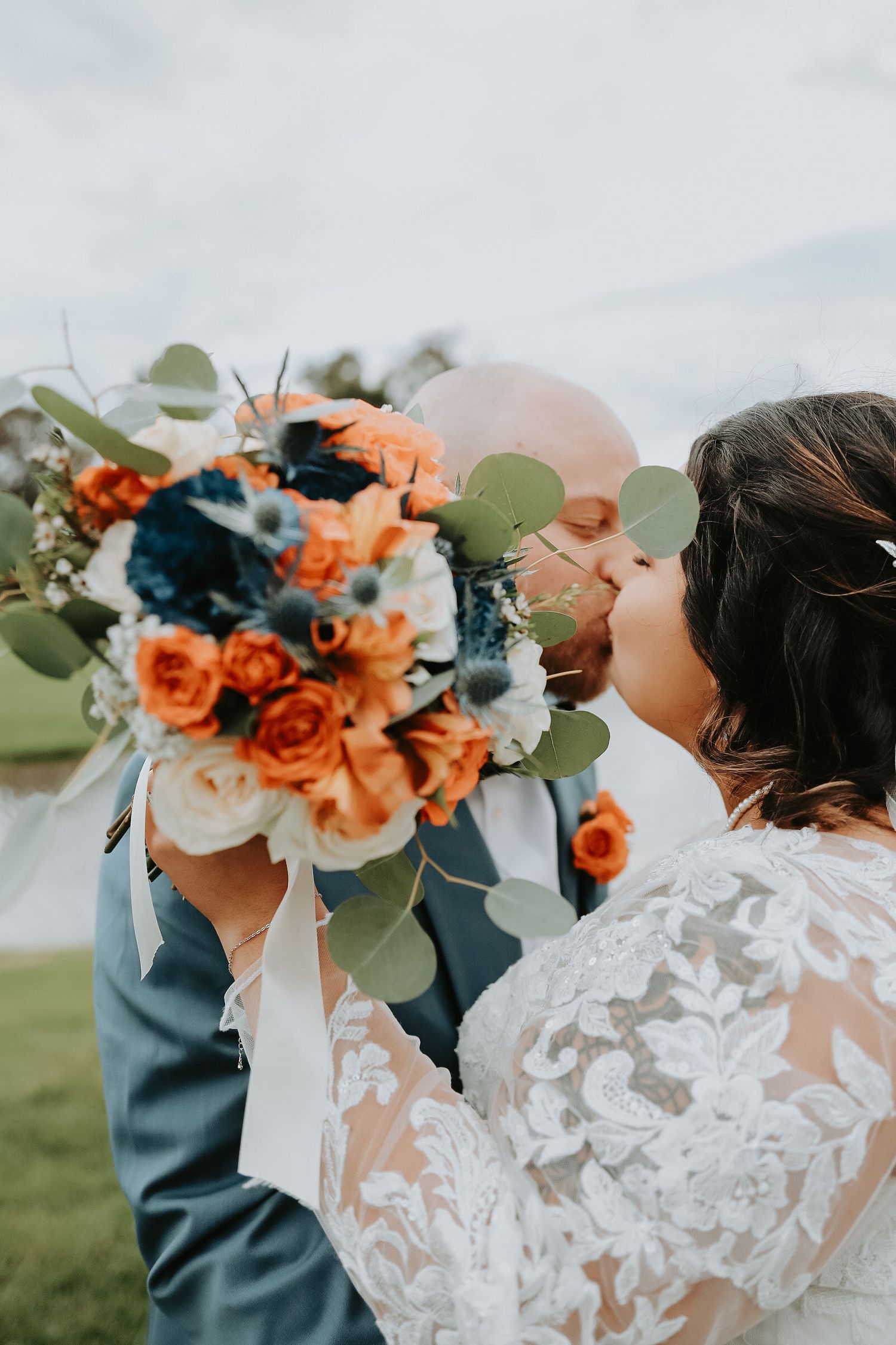 Wedding bouquet with orange roses, navy flowers and eucalyptus held against cloudy sky.