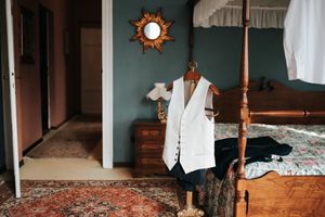 White groom vest hanging on a wooden four-poster bed in a vintage-styled room with a sunburst mirror.