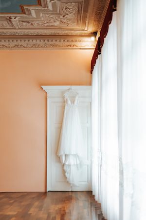 White lace wedding dress hanging on a wardrobe in an elegant room with ornate ceiling moldings and sheer curtains.
