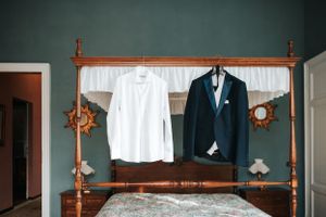 White dress shirt and navy blue groom blazer hanging on a rustic wooden four-poster bed with teal walls.