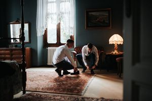 Two groomsmen helping each other with shoes in a vintage blue bedroom with warm lamp light and a Persian rug.