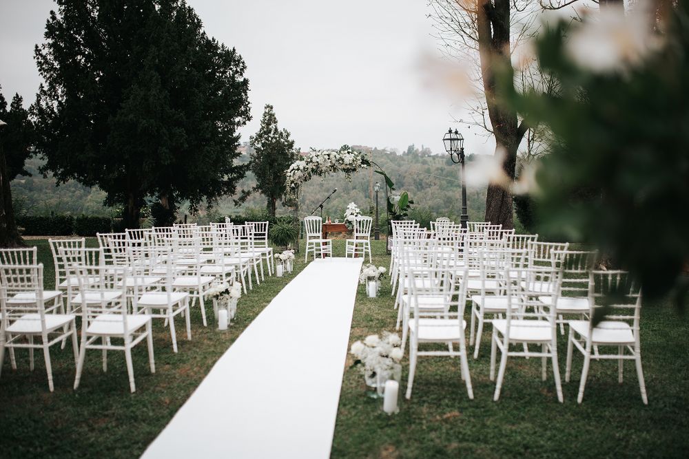 Outdoor wedding ceremony setup with white Chiavari chairs, white aisle runner, and floral arch in a garden.