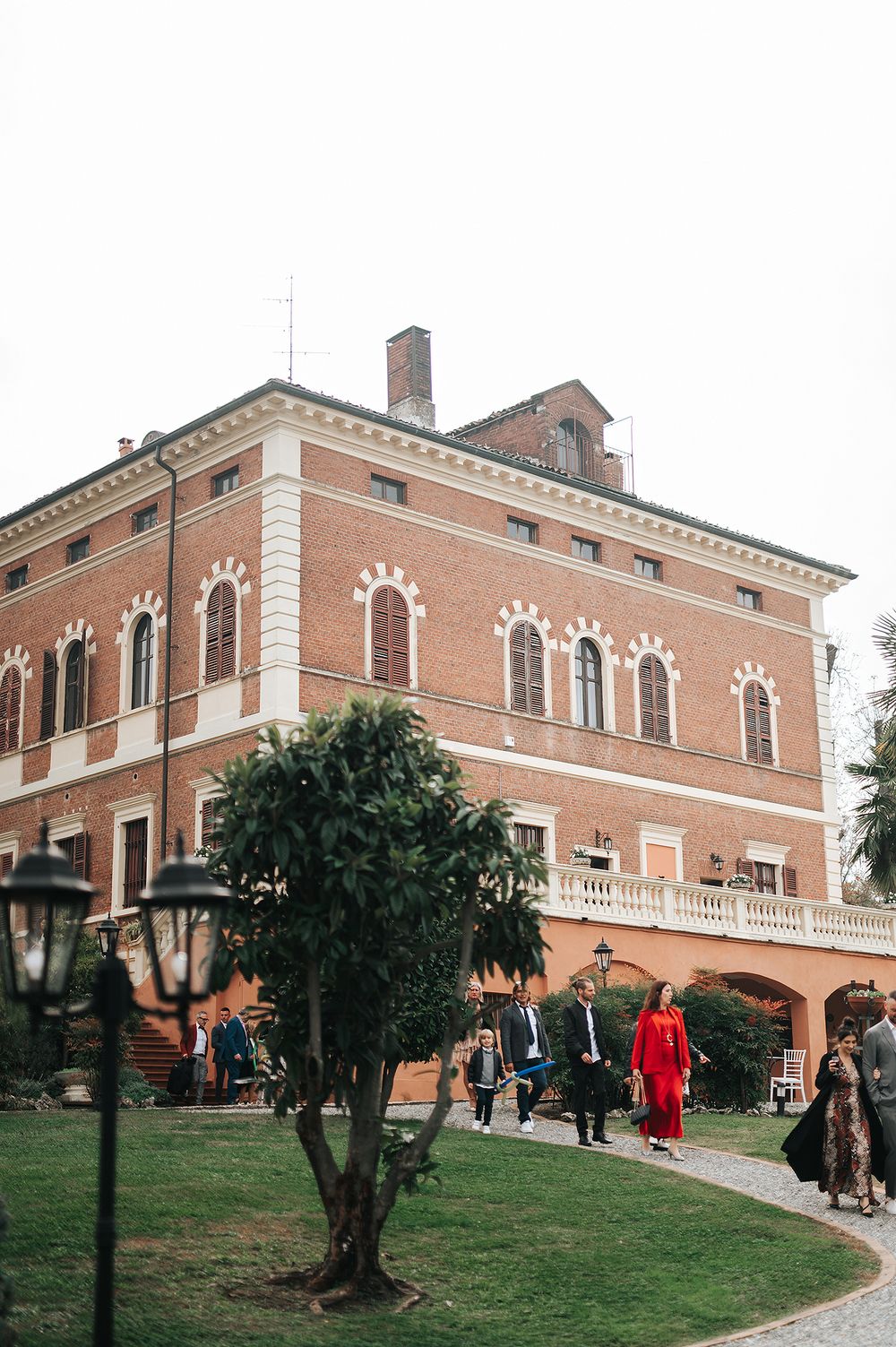 Elegant red brick Italian villa wedding venue with white balcony, ornate windows, and guests arriving on a gravel path.