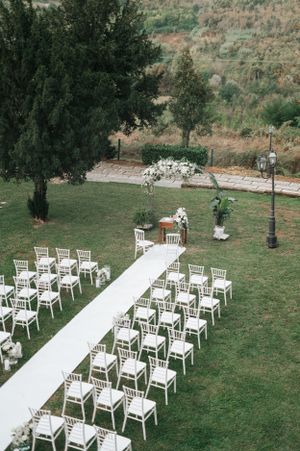 Aerial view of an outdoor wedding ceremony with white chairs, white aisle runner, and floral arch on green lawn.