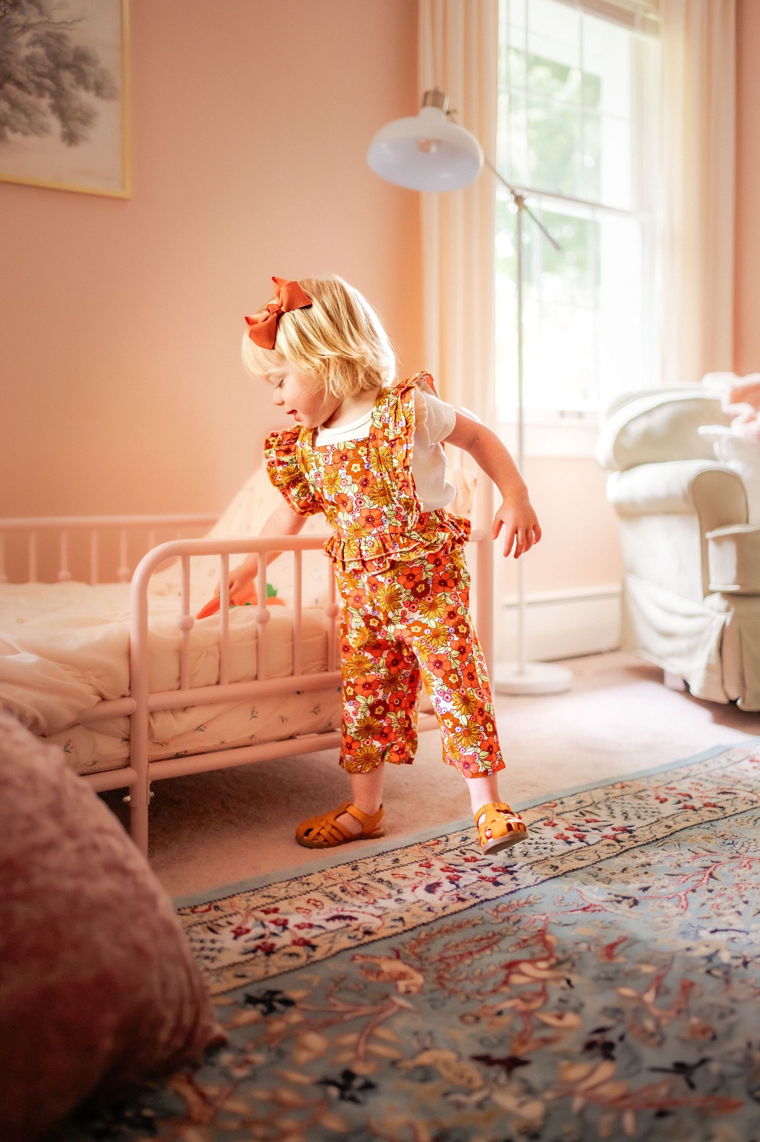 A vintage-styled nursery with pink walls features a decorative area rug and antique white metal bed frame.