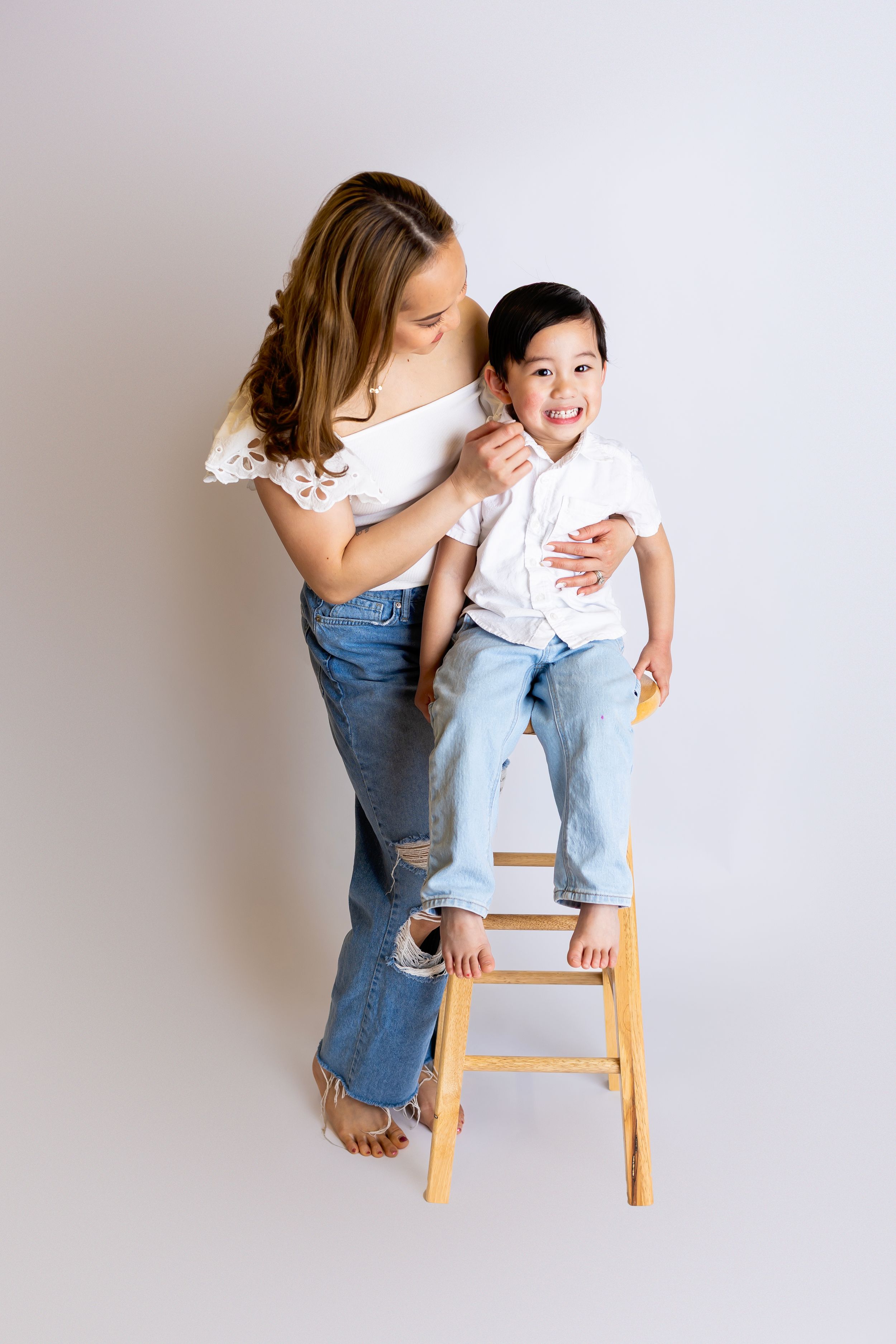 Two people in casual white tops and jeans share a playful moment on a wooden stool against a light background.