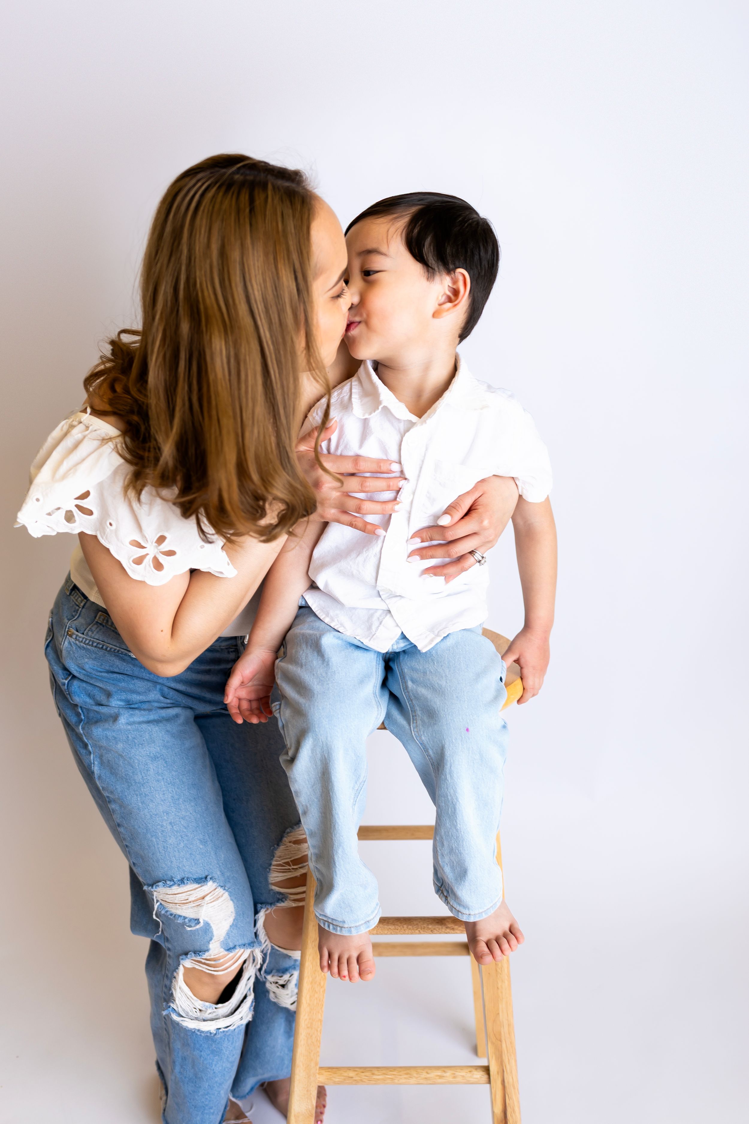 A tender moment shared between two people posed on a wooden stool against a white wall, wearing casual attire.