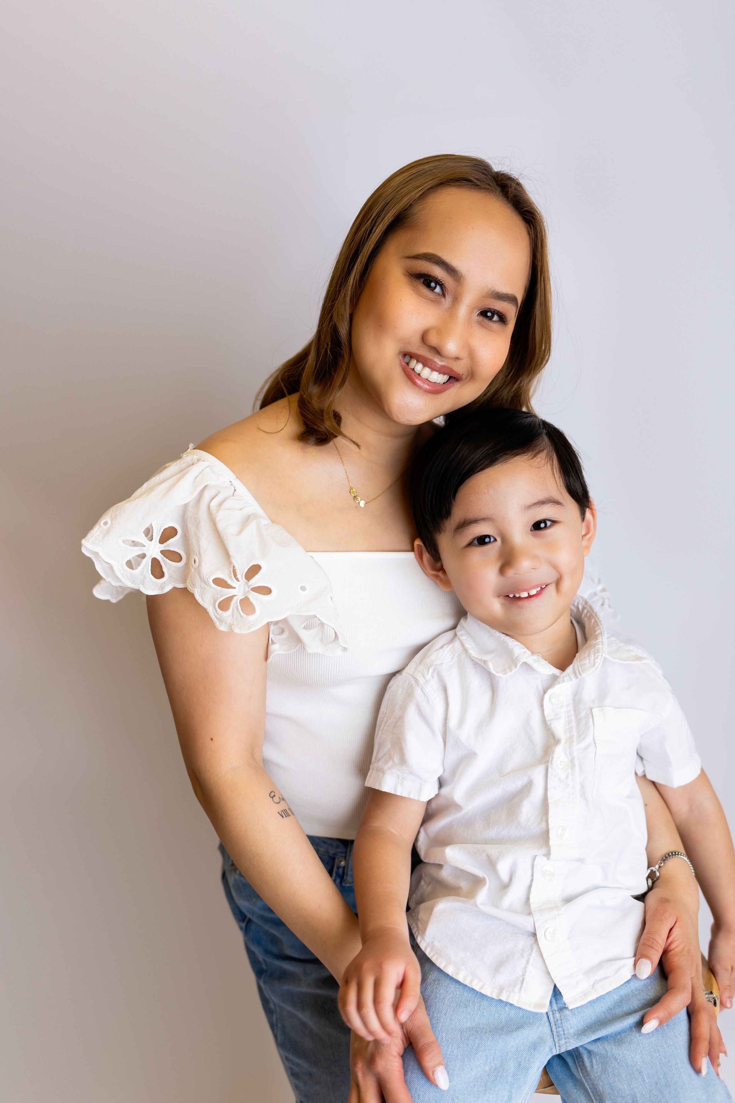 A young mother in a white eyelet top poses with her smiling son in a matching white button-down shirt against a gray wall.