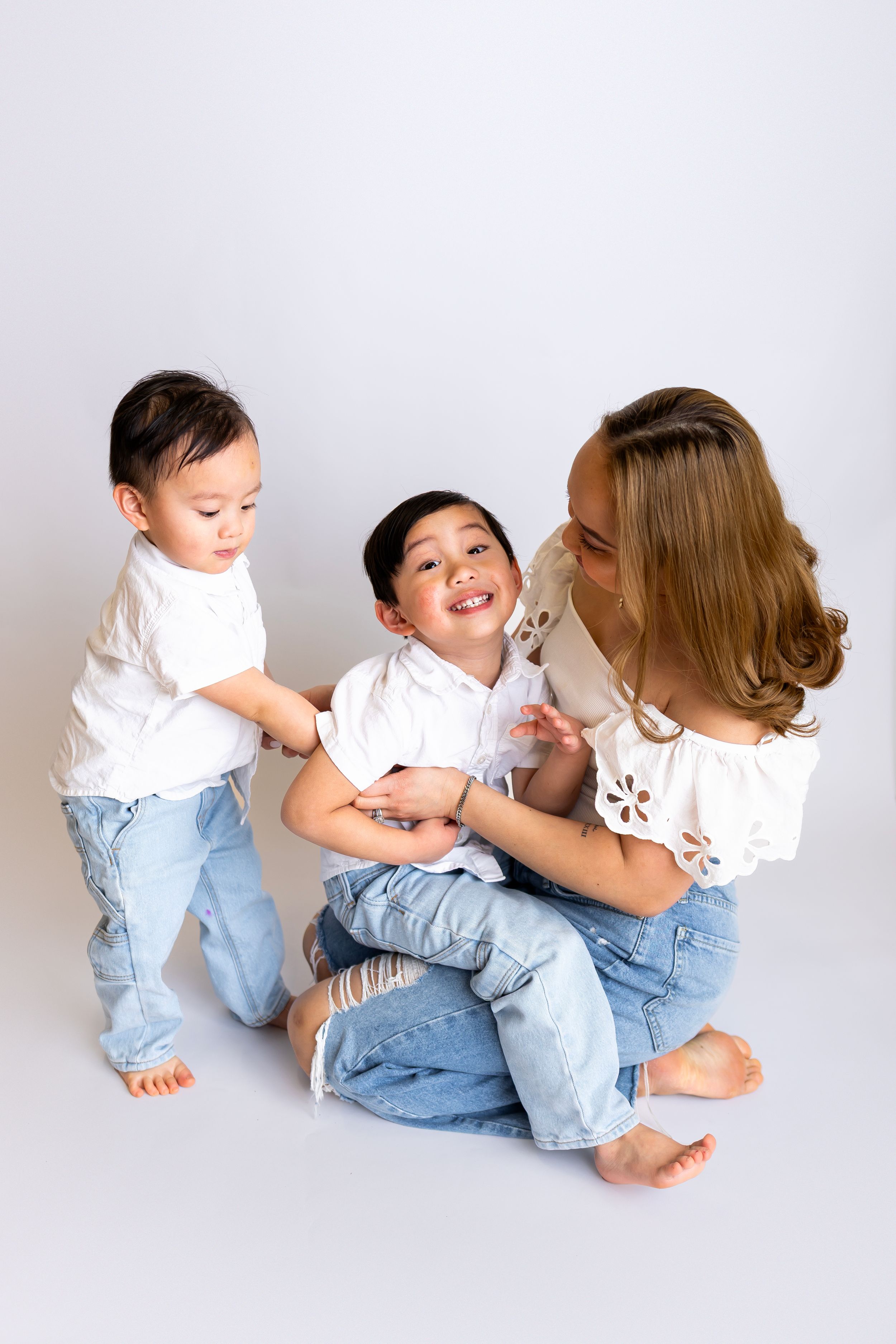 Three people wearing white tops and blue jeans sit and play together on a light gray background in a studio setting.