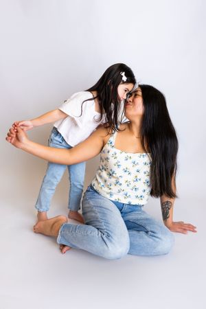 Two people with long dark hair share an affectionate playful moment wearing jeans and casual tops against a light background.