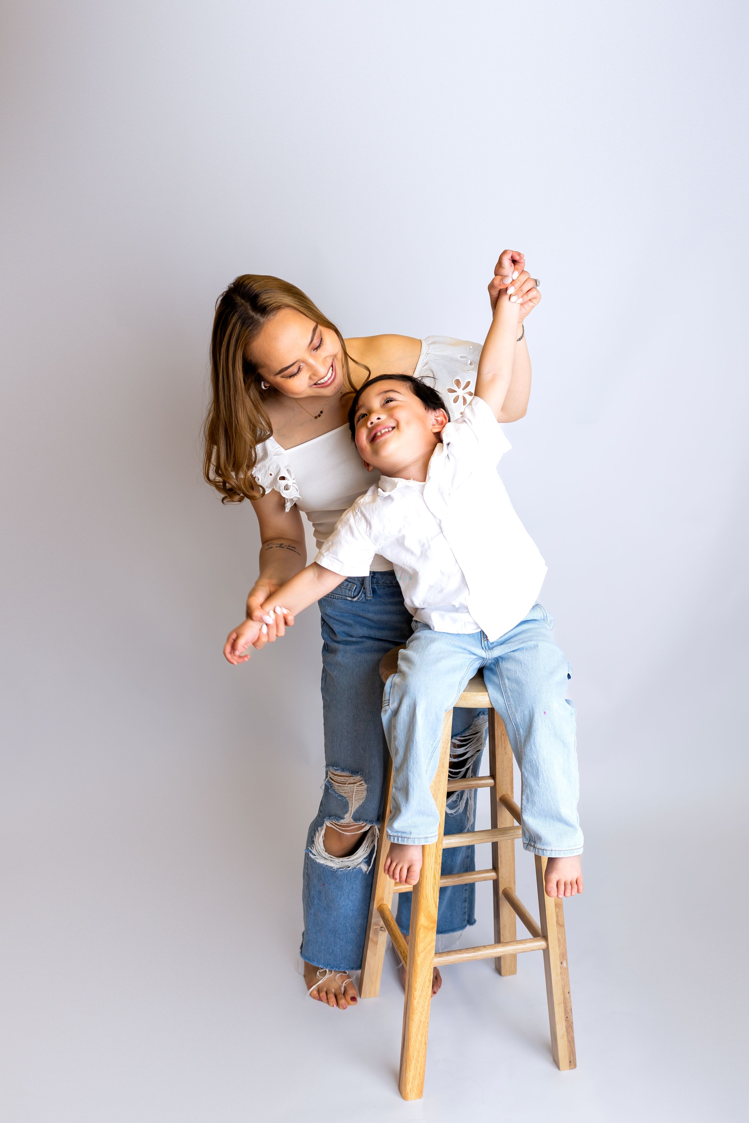 Playful moment captured with two people posing on wooden stool against light gray backdrop.