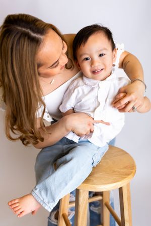 Joyful interaction between parent and child sitting together on a wooden stool against light background.