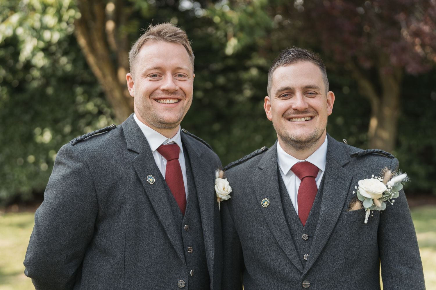 Two men in matching gray suits with red ties and white boutonnieres smile outdoors in a garden setting.