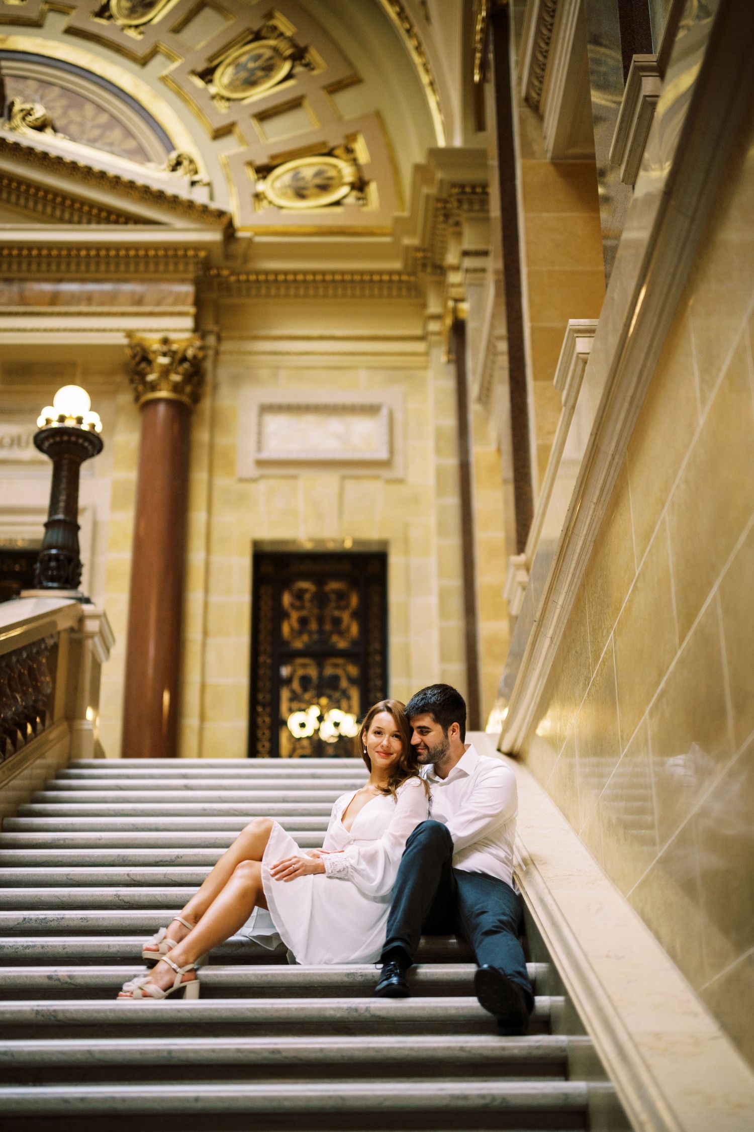 Couple sitting intimately on grand marble staircase inside ornate historic building with architectural details.