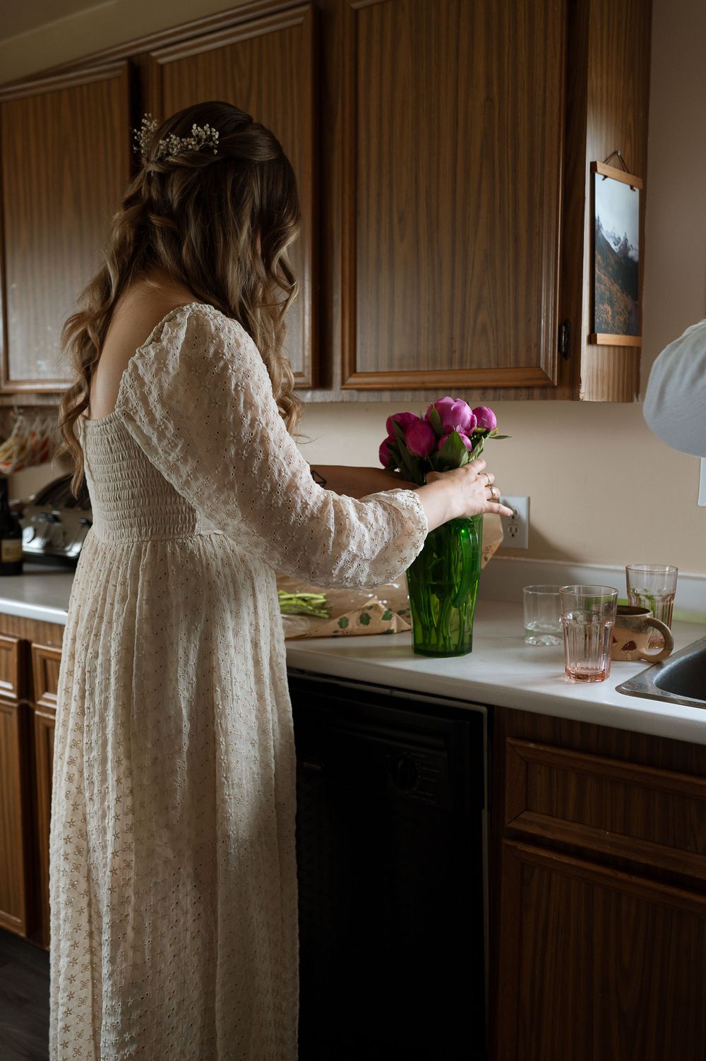 Person in white lace dress arranging flowers in green vase on kitchen counter.