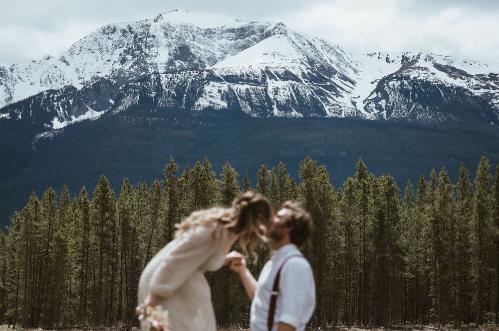 A romantic wedding couple embraces against a backdrop of snow-capped mountains and evergreen forest.