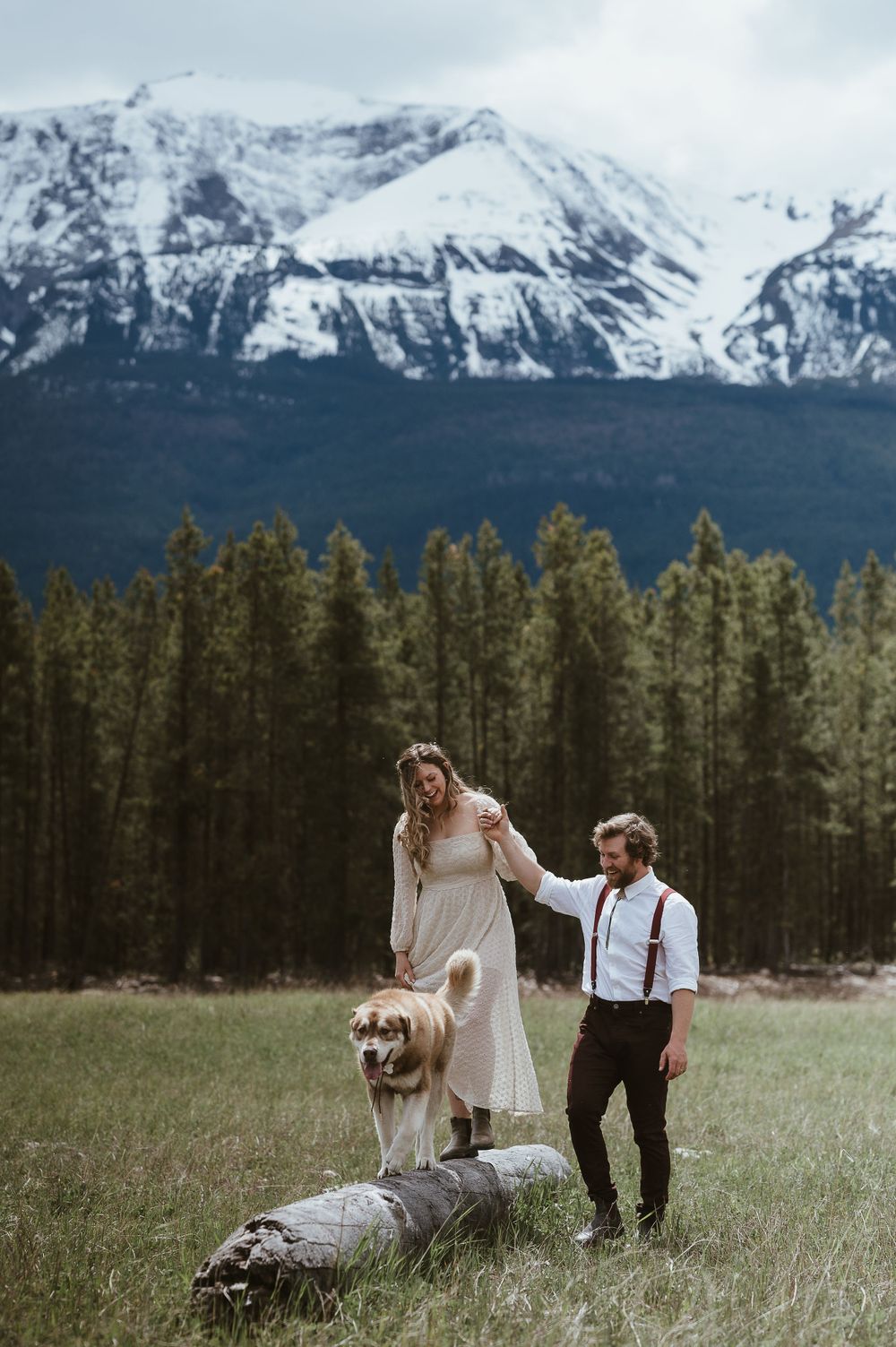 A couple and their dog pose together on a fallen log with majestic mountain peaks in the background.