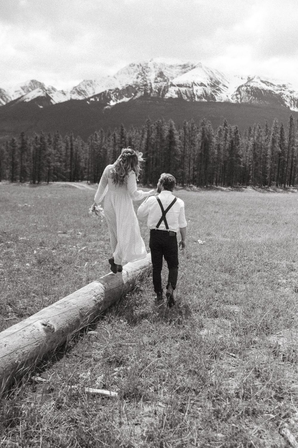 A couple walks along a fallen log in a mountain meadow, captured in black and white.