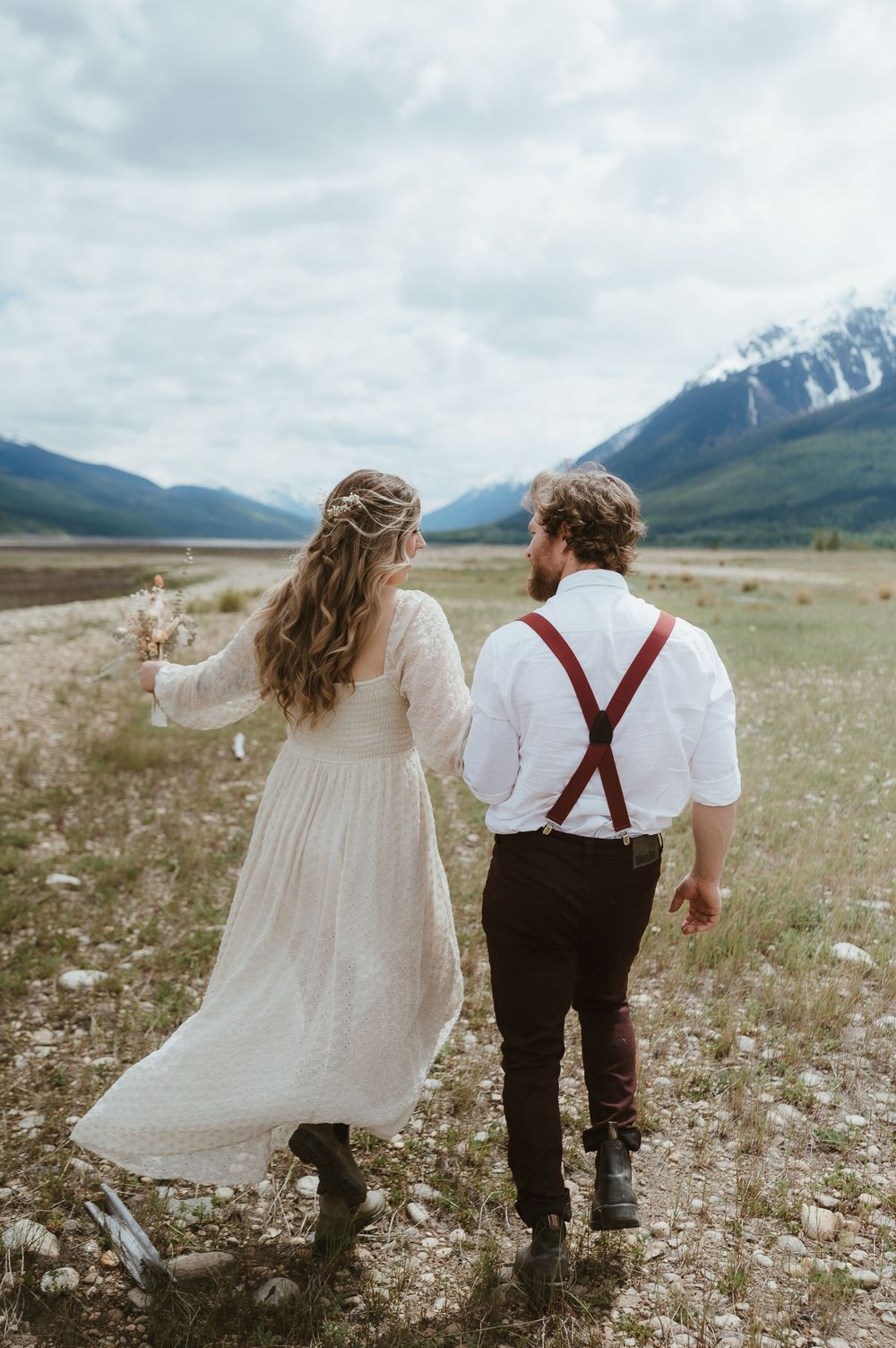 A couple walks hand in hand through a wildflower meadow with mountain views.