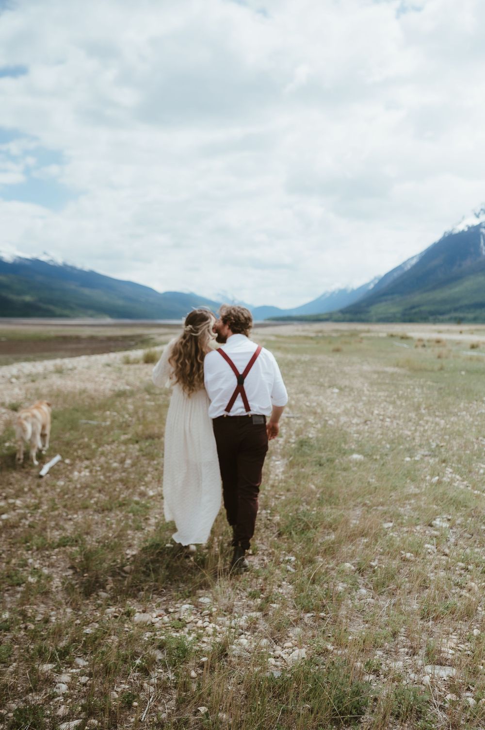 A couple walks through a mountain valley with their dog following behind.
