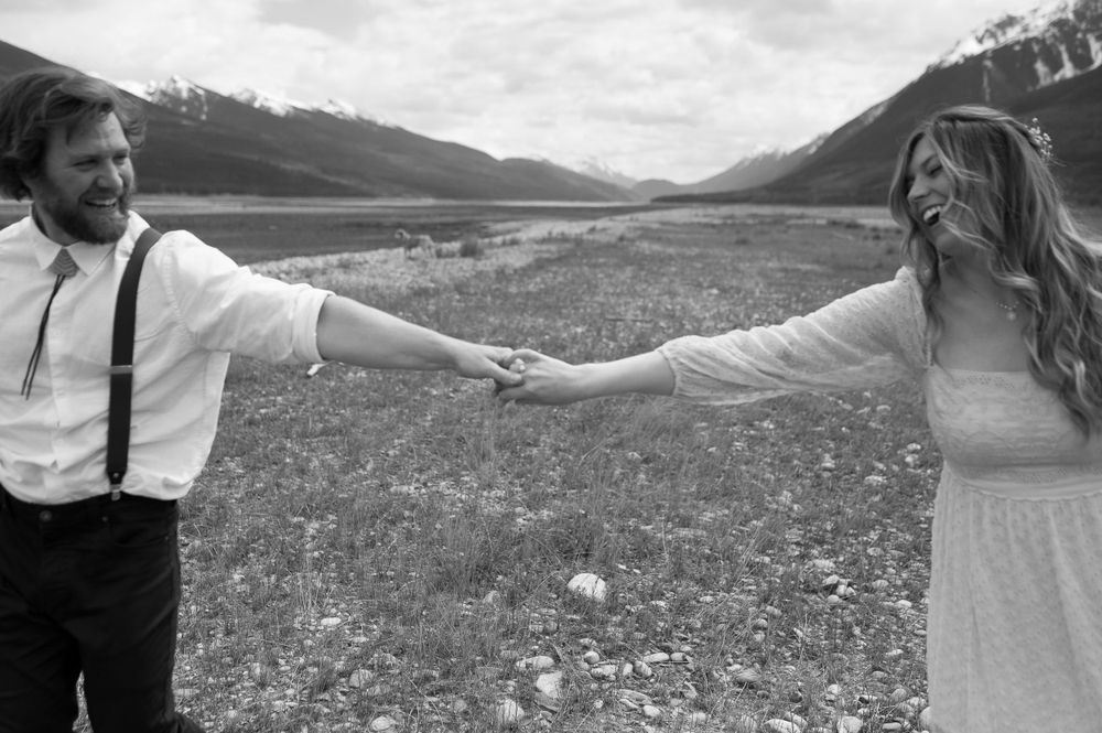 A romantic couple holds hands in a mountain meadow wearing formal attire against a backdrop of snow-capped peaks.