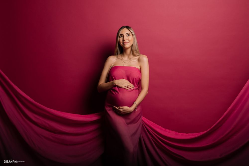 Maternity photo of figure in flowing deep red fabric against burgundy backdrop with dramatic drapery.