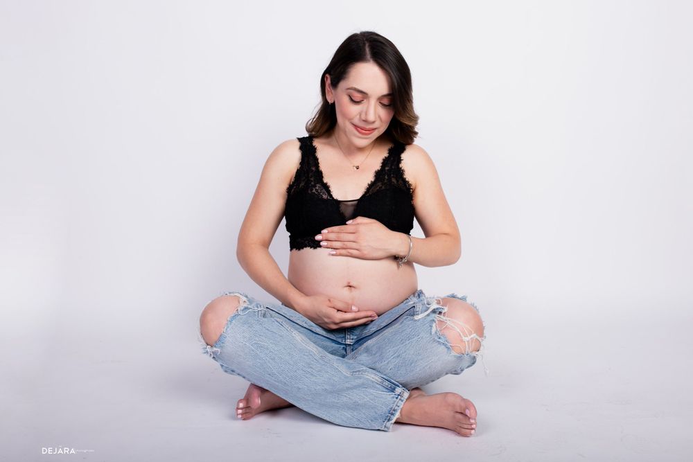 An expectant mother in casual denim sits cross-legged in a minimalist studio setting.
