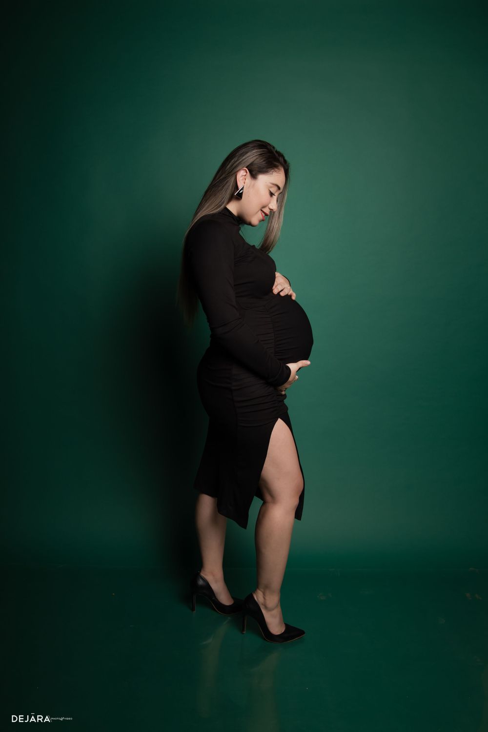 Dramatic maternity portrait in black dress against dark green studio backdrop.