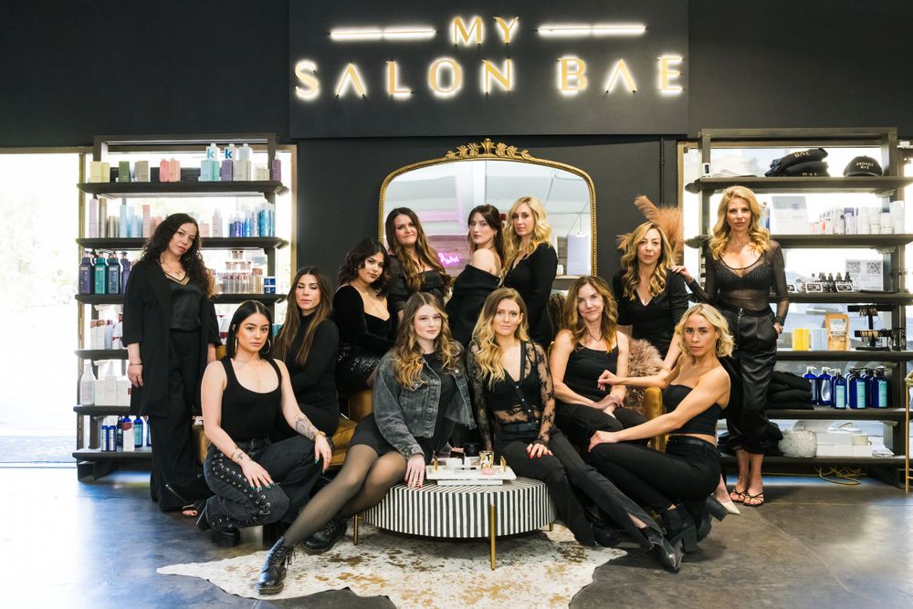 A group of salon professionals pose together in black attire inside a modern salon with My Salon Bae signage.