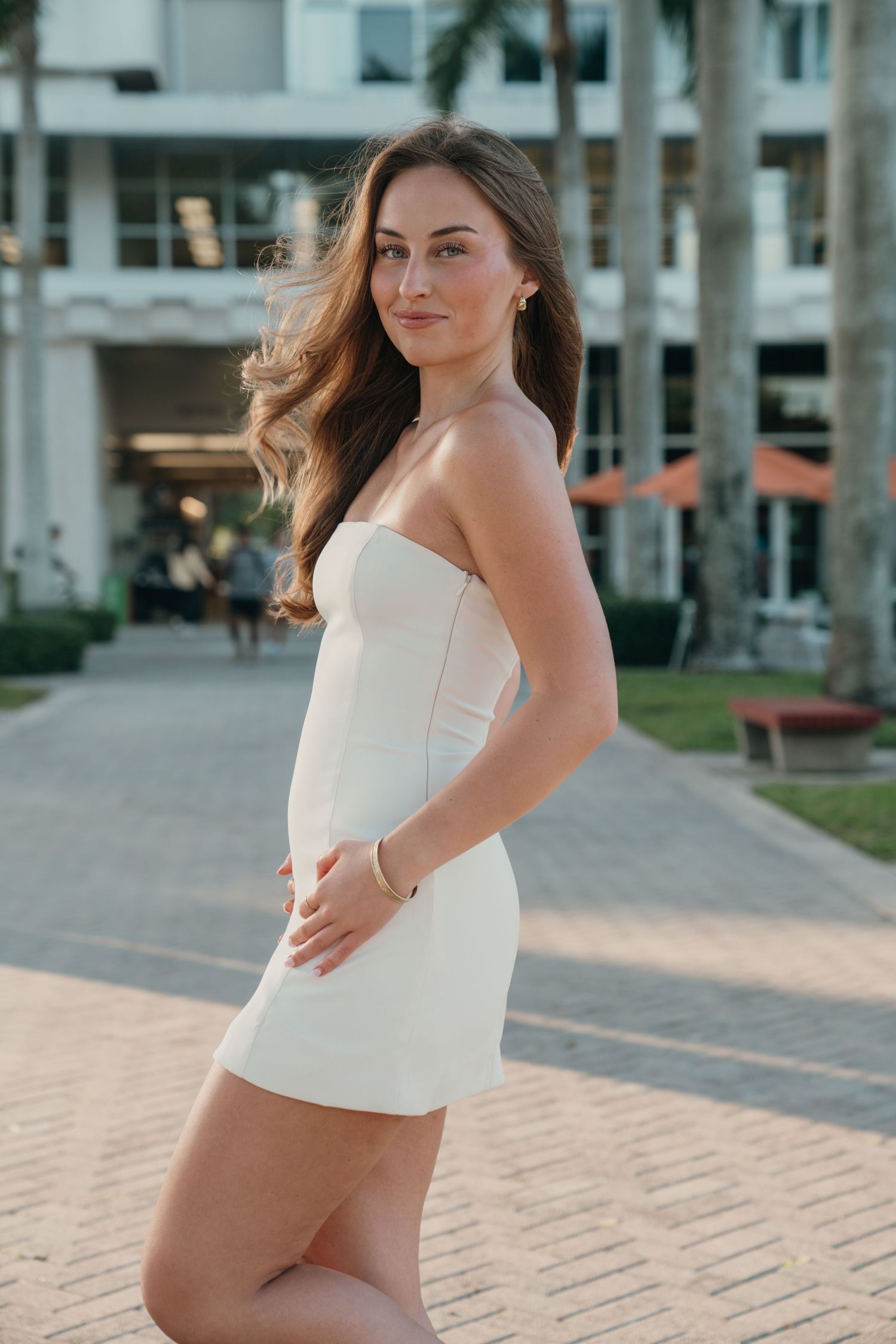 Person in white strapless mini dress poses outdoors at sunset near modern buildings and palm trees.