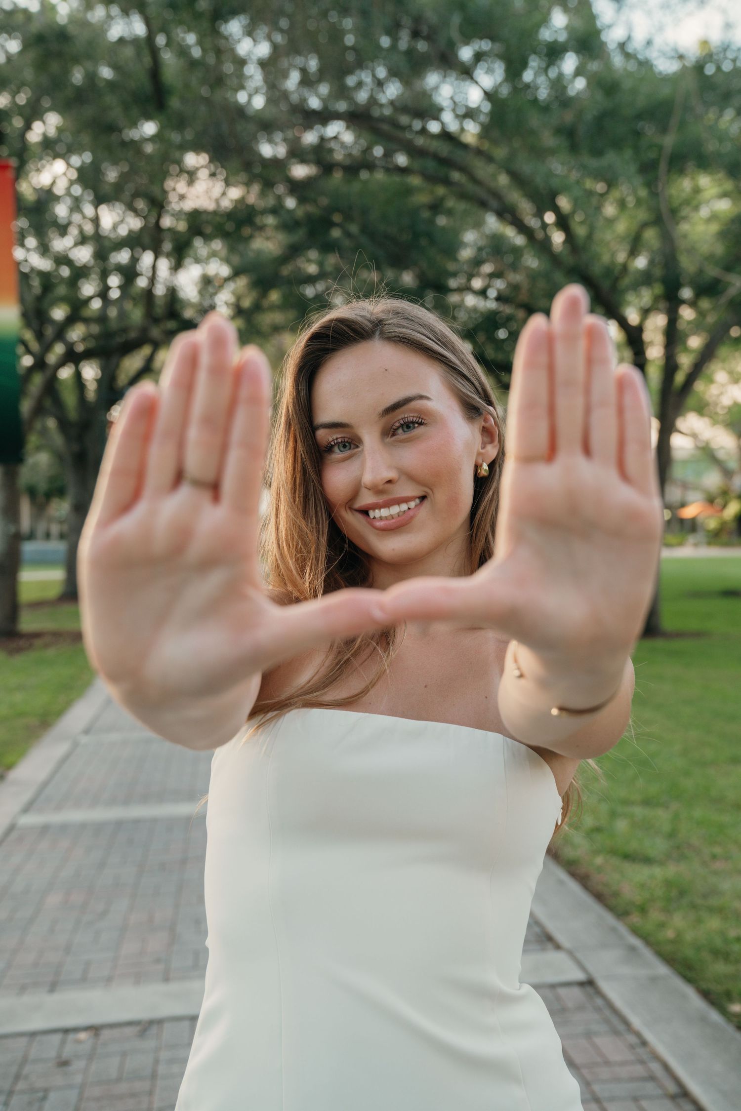 Person in white strapless top holds both hands up to camera while standing on brick path in park setting.