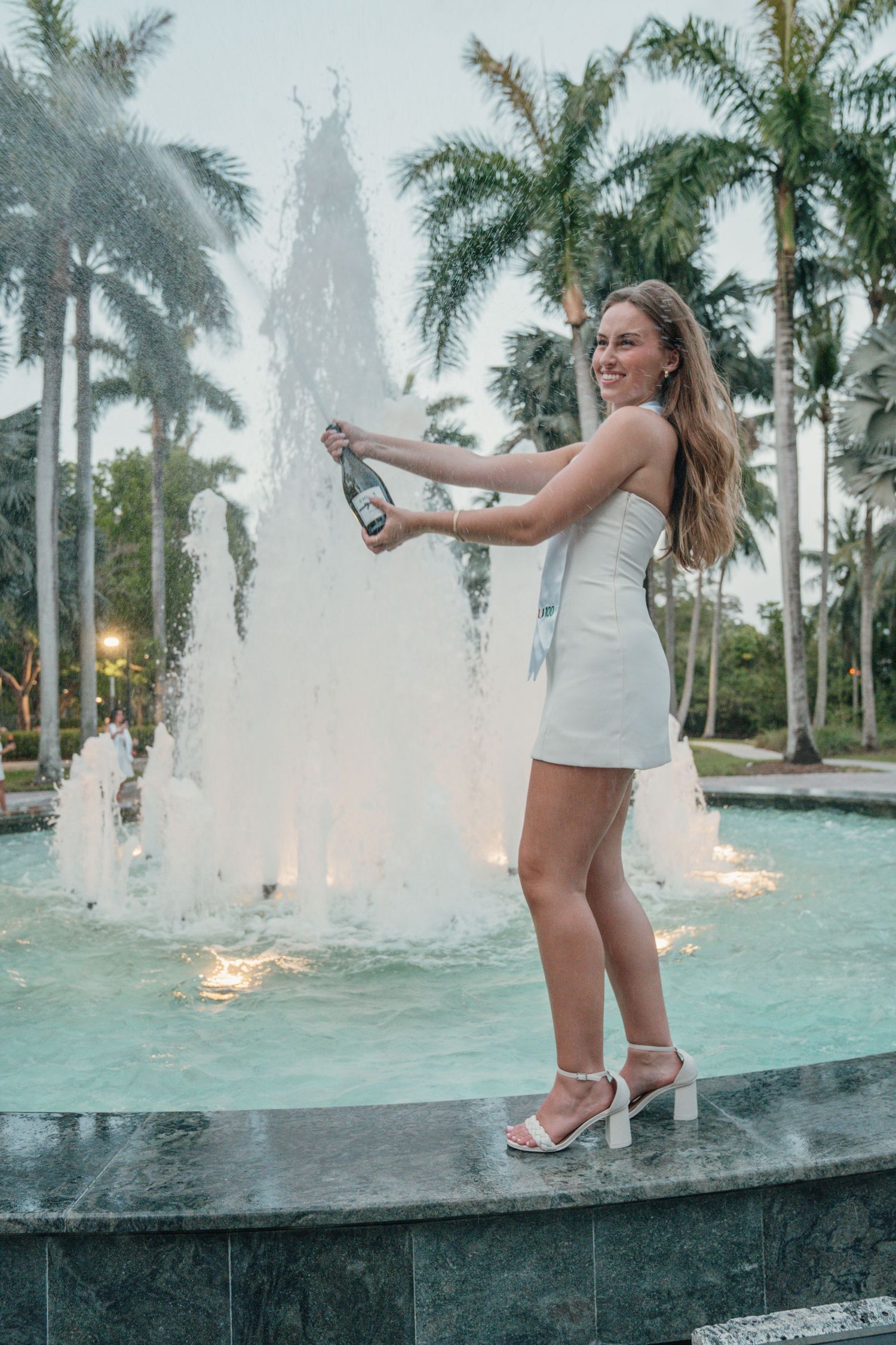 A woman in a white dress celebrates with champagne by a tropical fountain with illuminated palm trees at dusk.