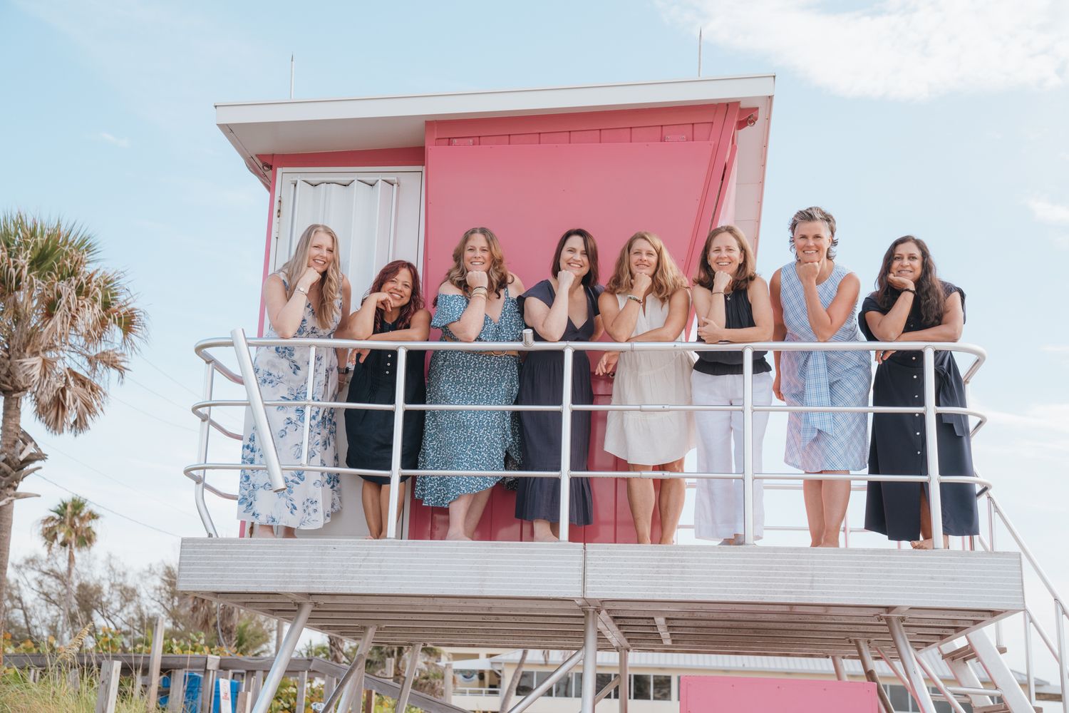 A group poses together on a pink and white beach lifeguard tower during a sunny oceanside photoshoot session.