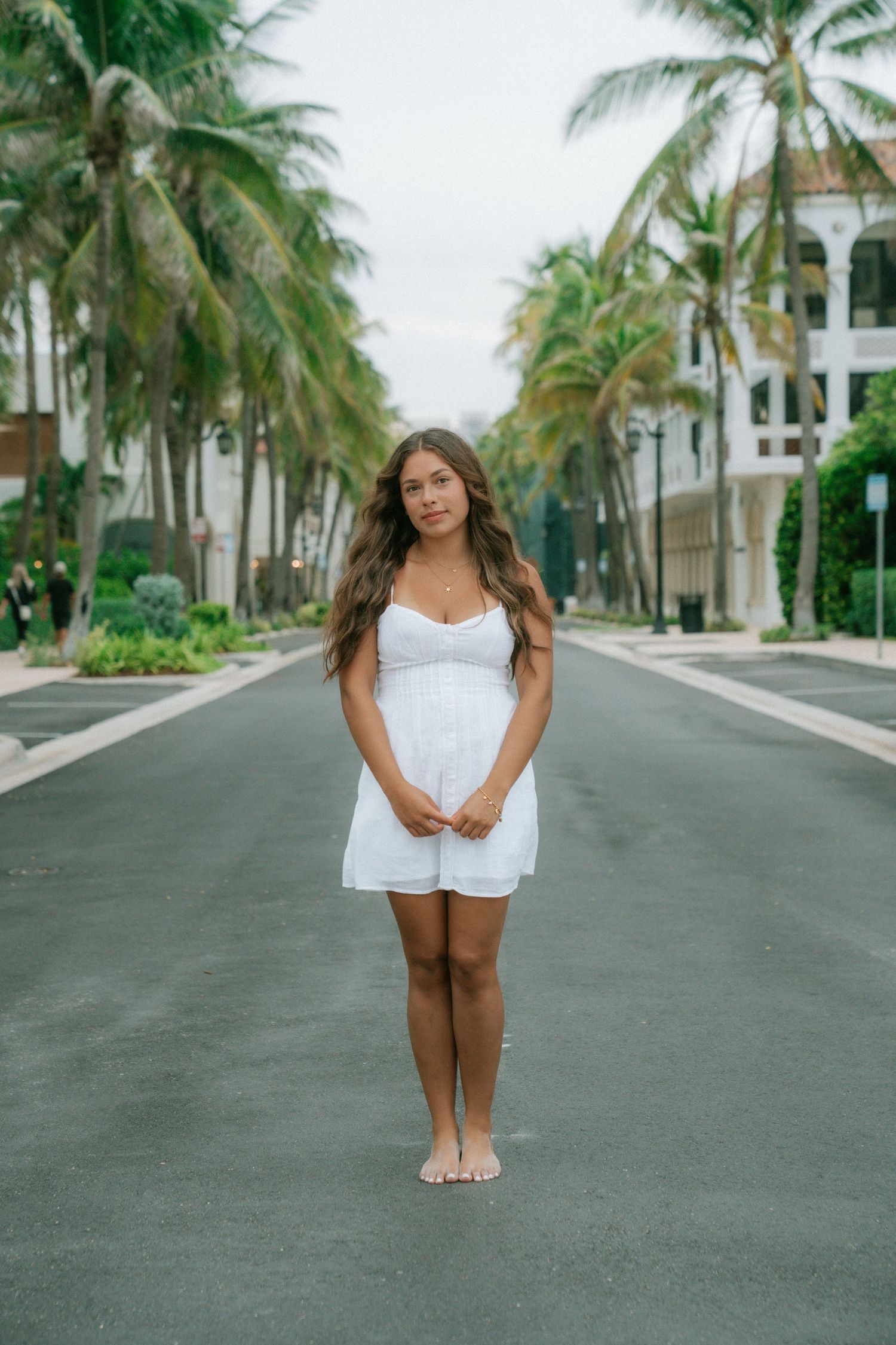 Young woman in white sundress standing barefoot on a palm-lined road at a tropical resort.