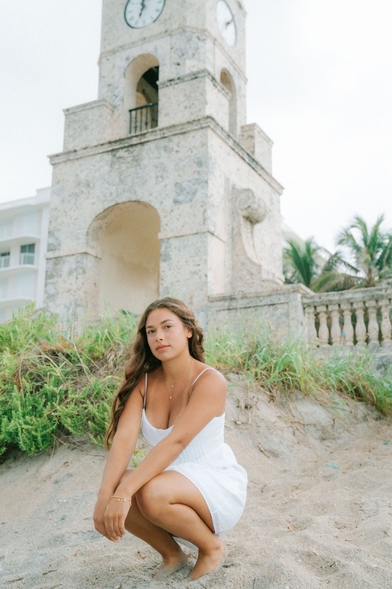 Young woman in white dress crouching on sandy beach near historic stone clock tower with palm trees.