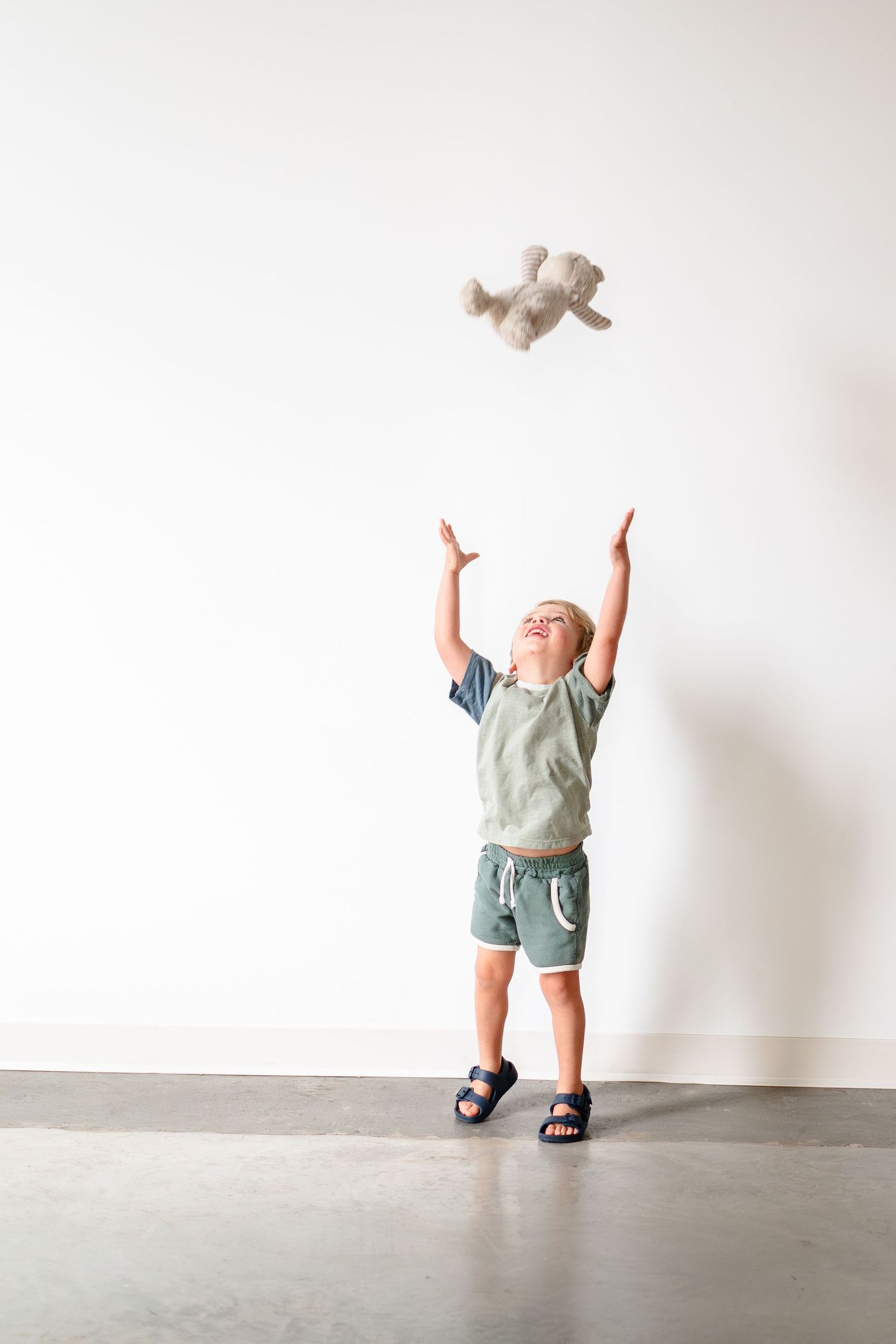 Child in sage green shirt and shorts joyfully tosses plush toy in the air against white wall.