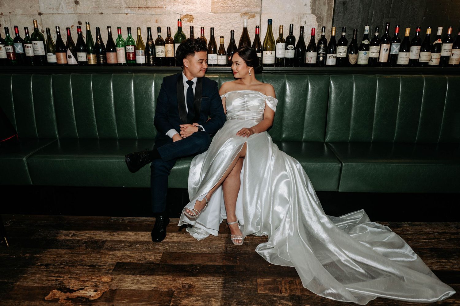 A couple poses on leather seating against a wall of wine bottles in formal wedding attire.